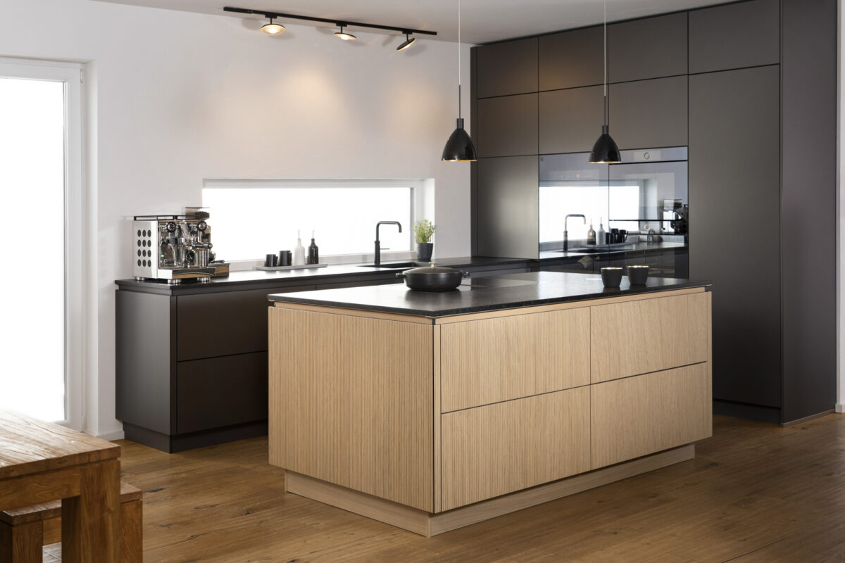 offene Designküche, zentrale Kücheninsel mit heller Holzfront, dunkle Hochschränke, großes Fenster und klare Linien. Interieur- und Küchenfotografie von Fotograf Robert Niedring in München