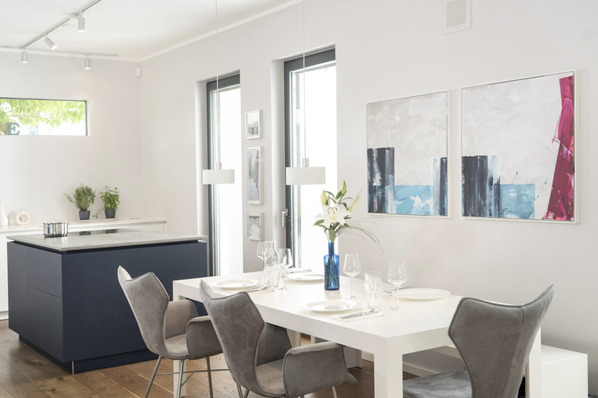 lichtdurchfluteter Wohn- und Essbereich mit weißem Tisch, modernen Stühlen, Kunst und Blick in den Küchenraum. Interieur- und Küchenfotografie von Fotograf Robert Niedring in München